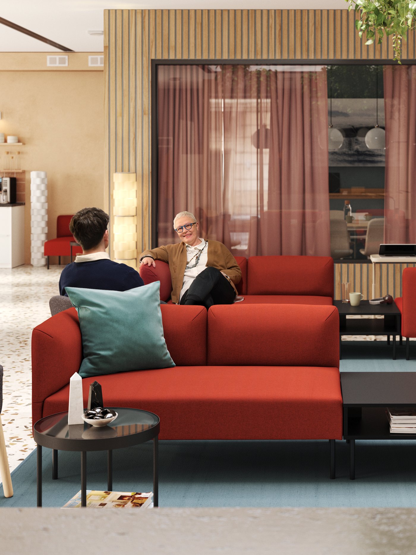 Two people relaxing in a modern, light-filled lobby and meeting room that has two red LILLEHEM modular sofas in the centre.