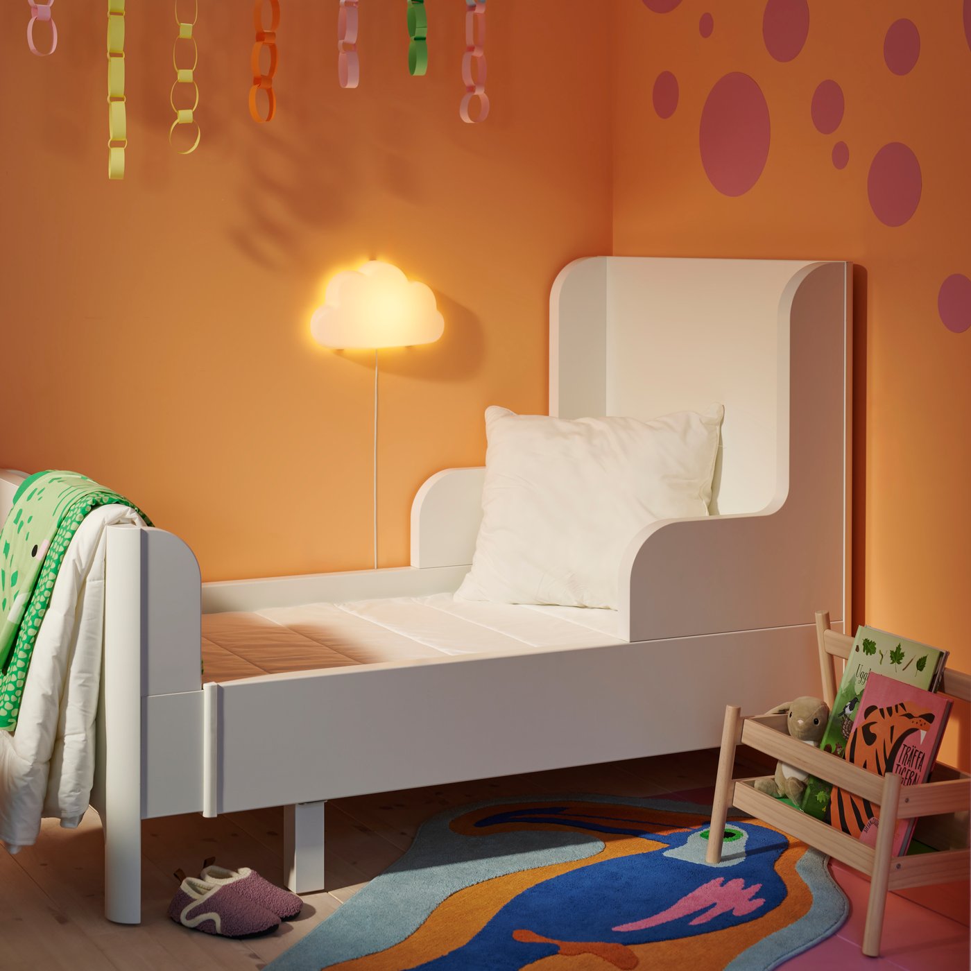 Modernes weißes Kinderbett in einem Zimmer mit orangefarbener Wand und runder Wandlampe. Bunte Teppiche und hängende Dekorationen sorgen für fröhliche Akzente.