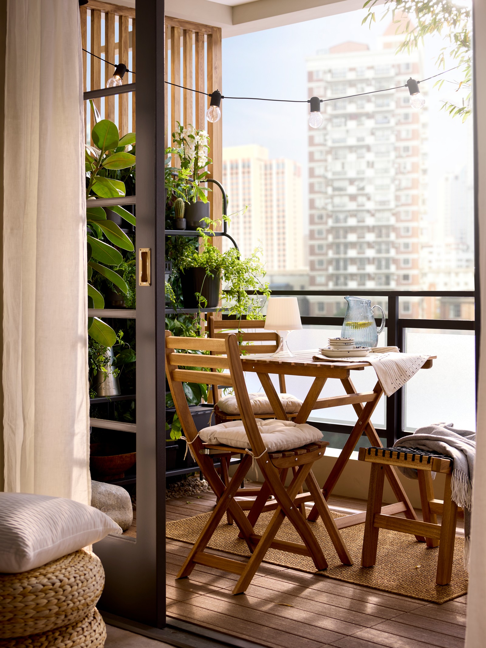 A balcony has a light brown stained acacia ASKHOLMEN foldable table and 2 folding chairs by potted plants on a side wall.