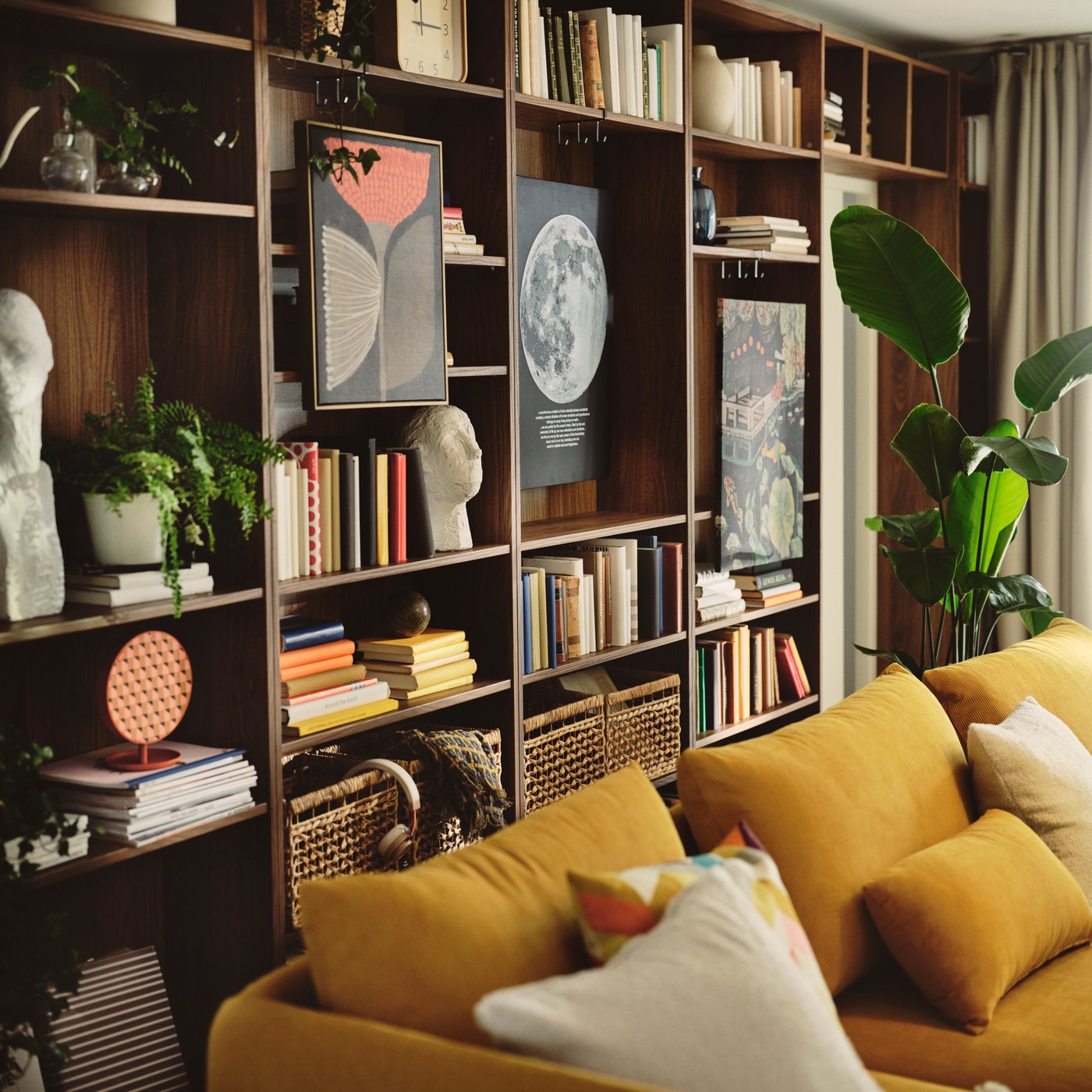 A living room with a brown BILLY bookcase in walnut effect, filled with books and decorative items near a yellow sofa.