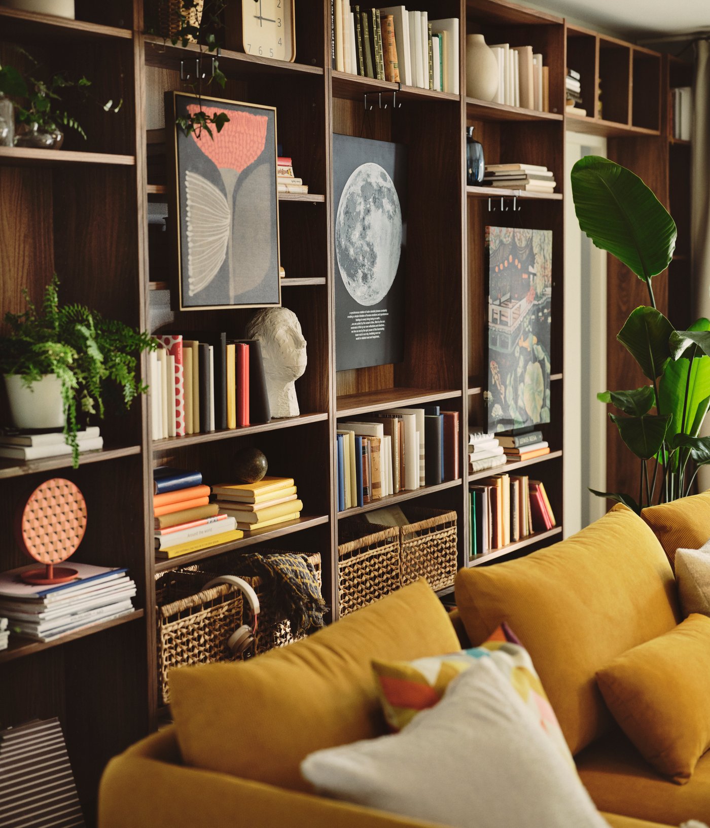 A living room with a brown BILLY bookcase in walnut effect, filled with books and decorative items near a yellow sofa.