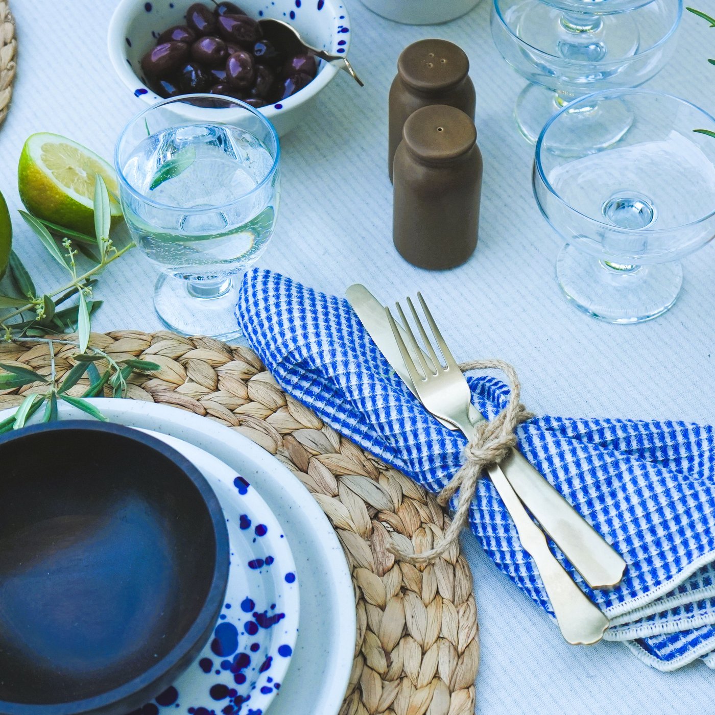 Une table dressée dans un style estival avec des couverts NATTKLOT, un bol d'olives, des citrons verts frais et un pot d'herbes aromatiques, idéale pour un repas méditerranéen.