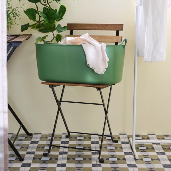 A big green plastic TORKIS flexi laundry basket is on a folding chair by a drying rack with pink fabric draped over its side.
