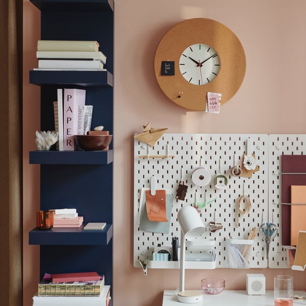 An ENSMÄRKE desk lamp stands on a white desk with SKÅDIS pegboards and a SVARTGÖK wall clock attached to the wall above.