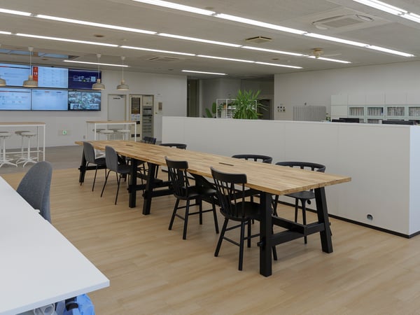 開放的なオフィスのワークスペース。木製テーブルと黒いチェアが並び、観葉植物と大きなモニターがある明るい職場環境。