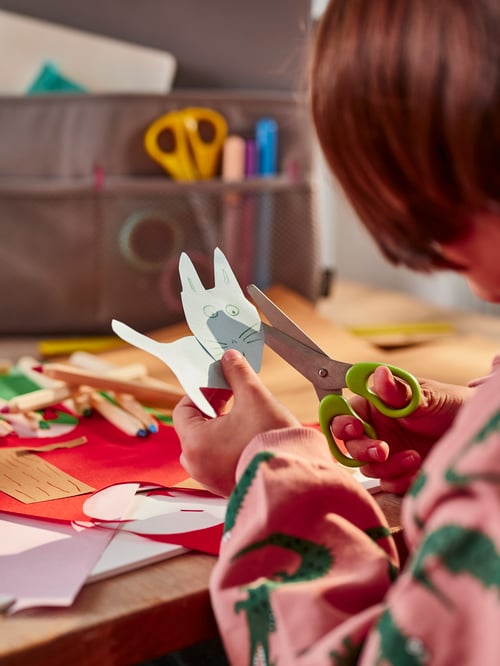 A child using MÅLA scissors to cut a paper cat. Scattered MÅLA mixed-colour paper lie on a nearby tabletop.