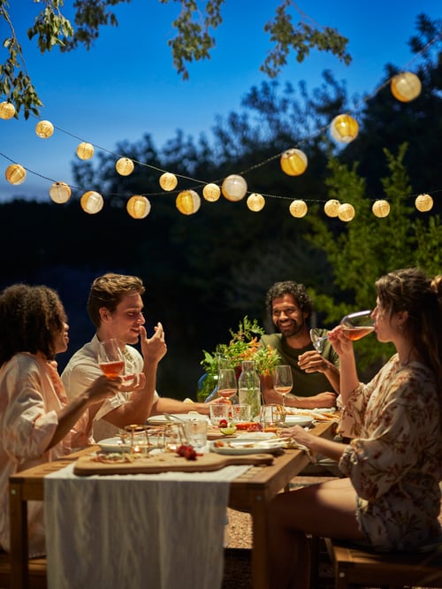 An outdoor setting featuring a lit SOMMARLÅNKE LED lighting chain with 12 lights placed above a table with people.