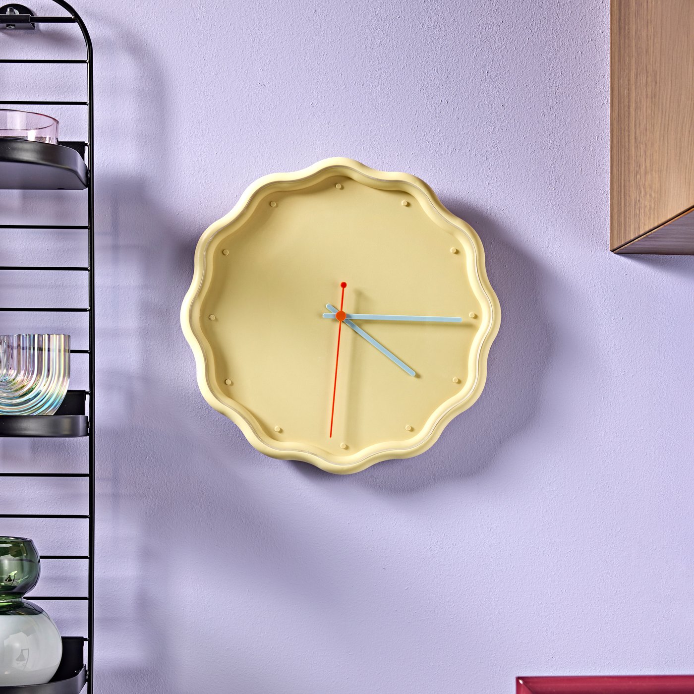 A close-up of a LAVSKRIKA clock, yellow with a scalloped edge, on a purple wall beside shelves with colourful glassware.