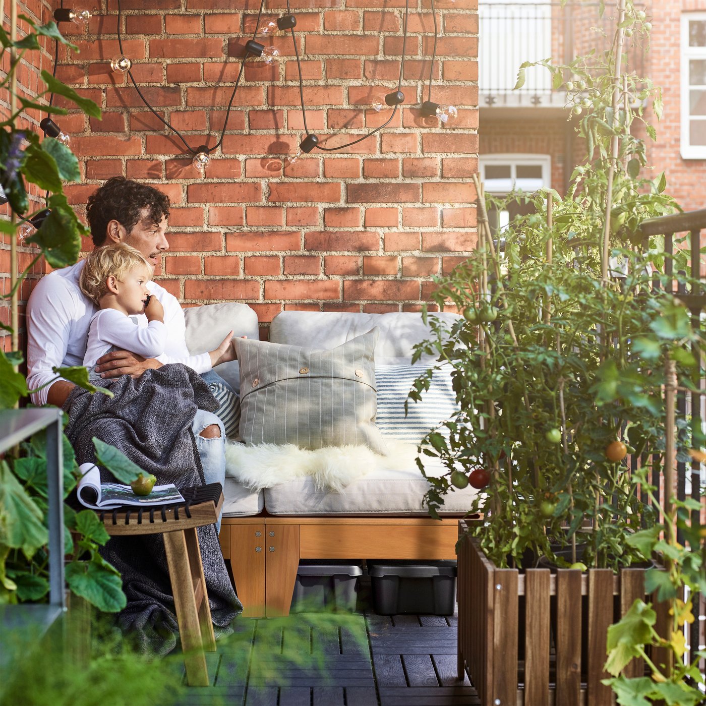 Ein Mann und ein Kind sitzen auf ÄPPLARÖ Sitzelement für draussen braun lasiert vor einer roten Backsteinwand auf einem Balkon.