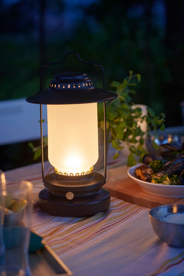 Vintage-style STORHAGA LED table lamp glowing warmly on a dinner table outdoors at night, surrounded by plates of food and leafy plants, creating a cozy ambience.
