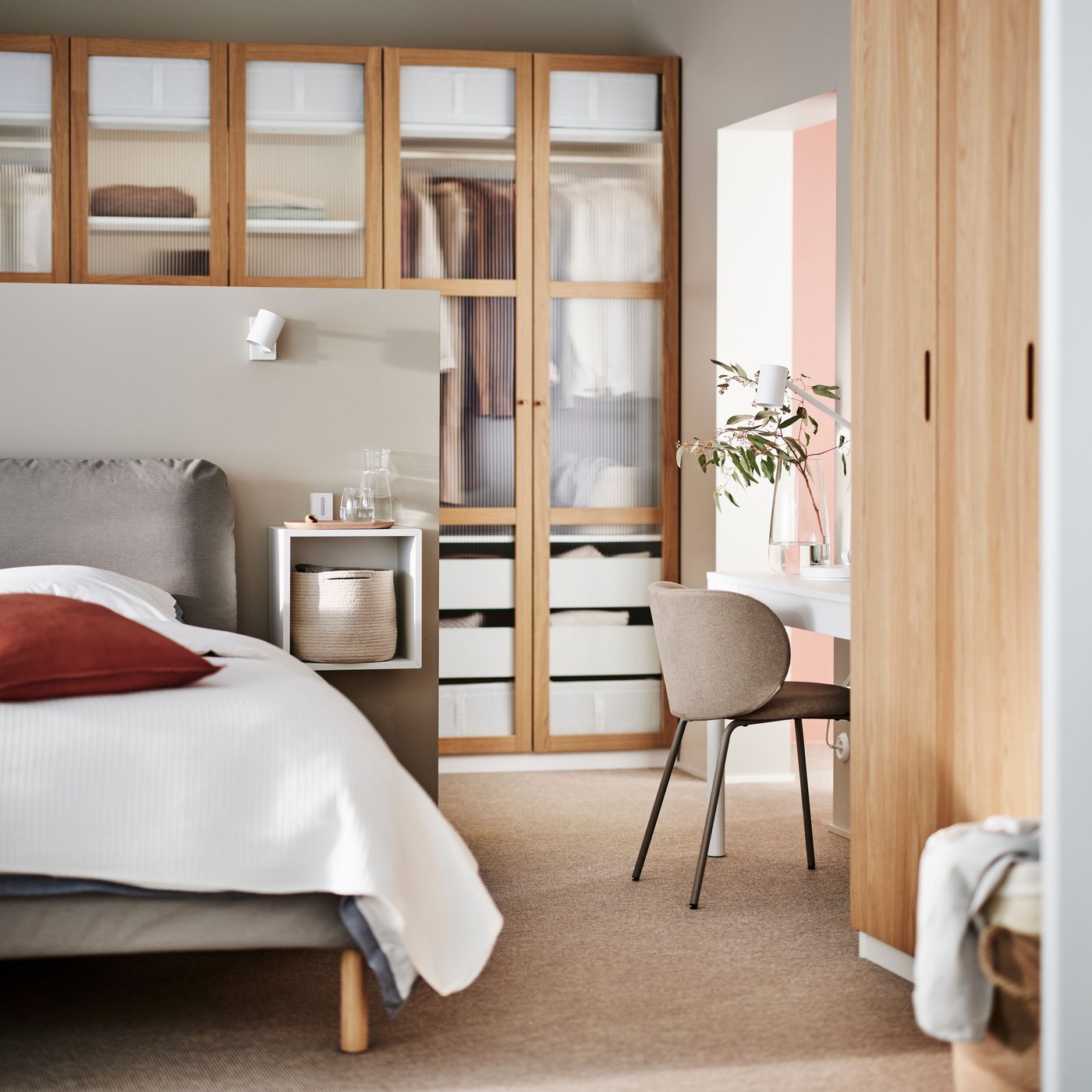 A brown SAGESUND upholstered bed covered in white bed linen in front of a PAX/TONSTAD wardrobe in white oak veneer/glass.