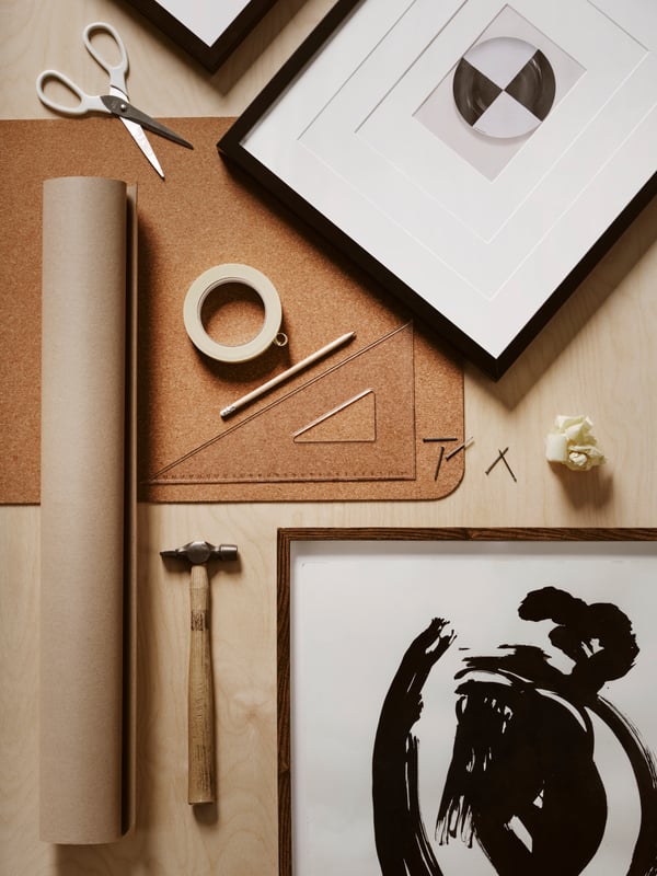 A desktop with RIBBA and LOMVIKEN frames, a roll of brown paper, a roll of tape, scissors, a hammer, a ruler and a pencil.