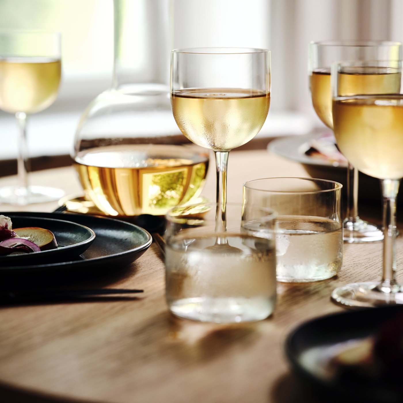 A table by a window is set with STOCKHOLM 2025 wine glasses and other glasses, a carafe, plates, and side plates.