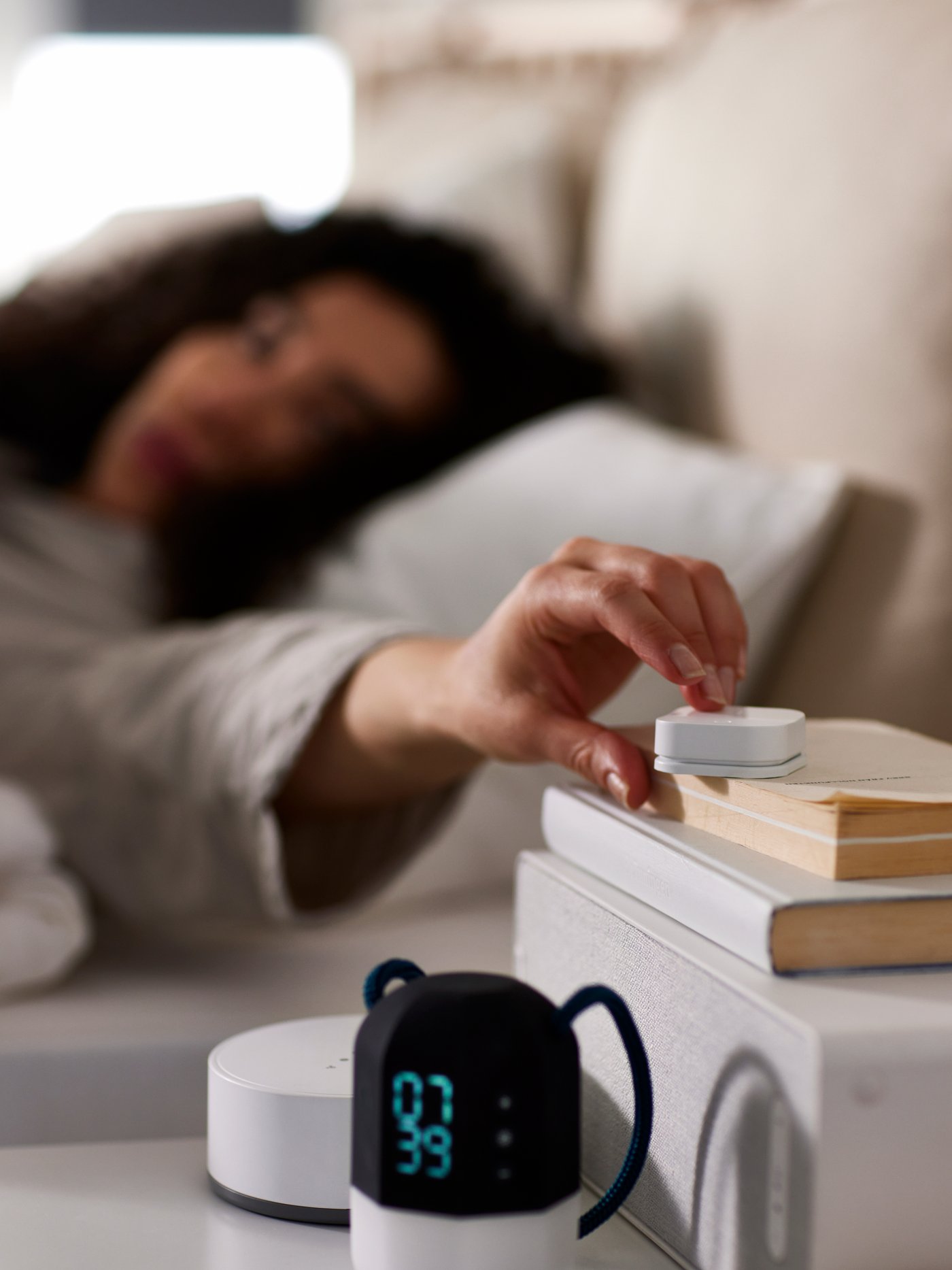 A woman lying in bed pushing a TRÅDFRI wireless dimmer on top of some books and a WiFi speaker on a white bedside table.