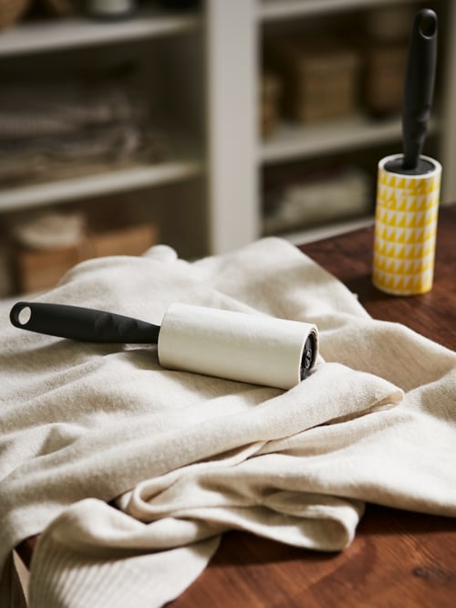 A wooden surface with a knitted, beige jumper, one BÄSTIS lint roller lying on it and a new lint roller standing next to it.