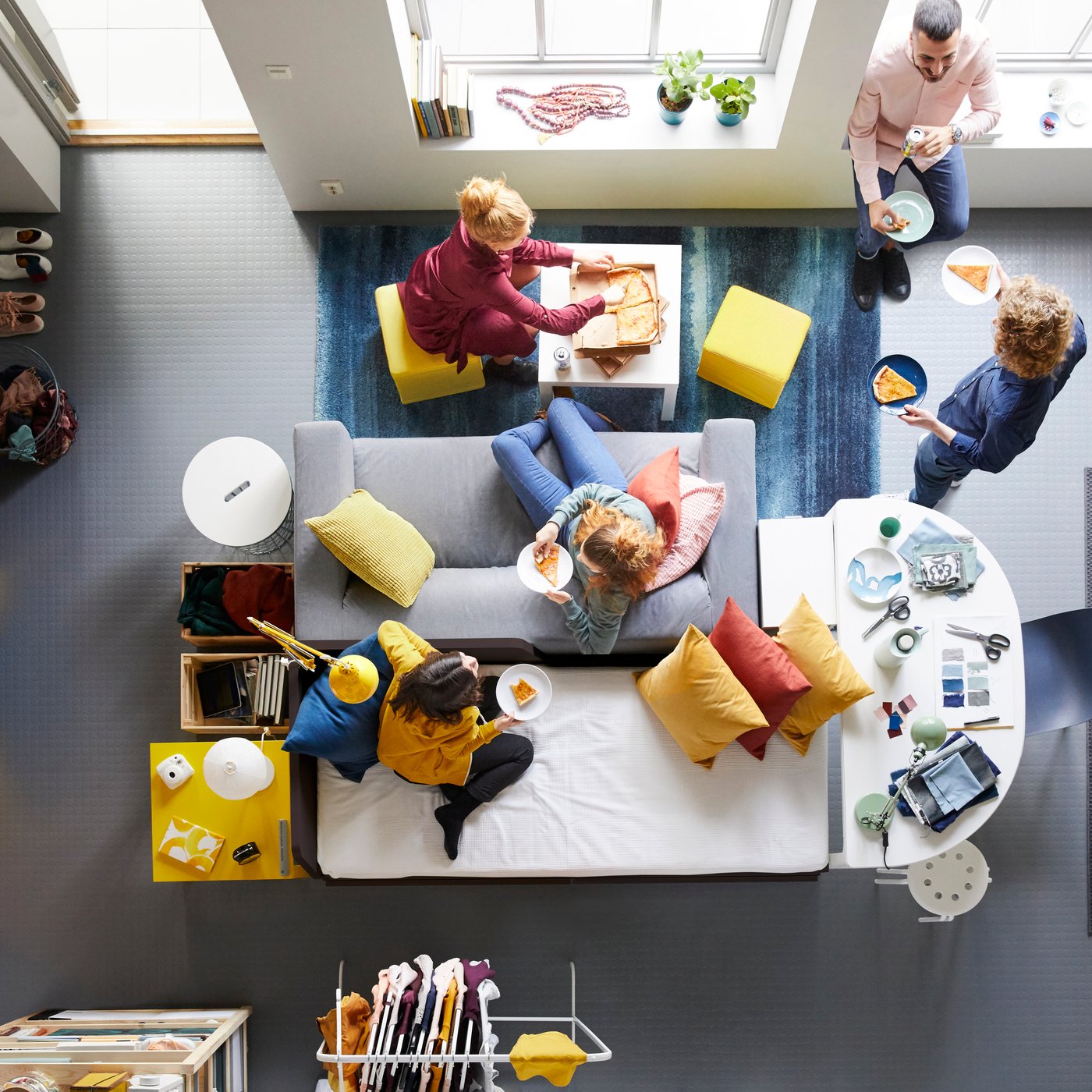 Plano panorâmico de sala com quatro pessoas a descontrair no e à volta do sofá KIVIK em cinzento.