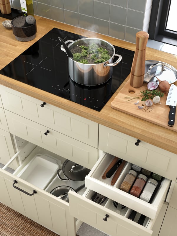 An IKEA 365+ pot with lid on a black induction hob, with open kitchen drawers below with pots, oven dishes and spices inside.