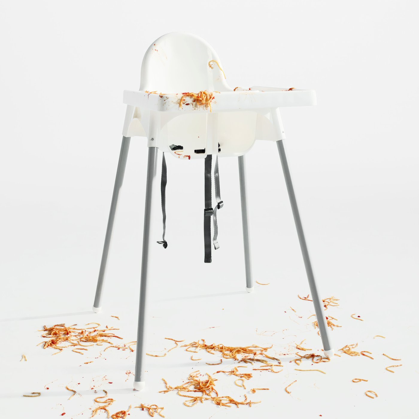 An empty highchair with tray in a white space. The remains of a messy spaghetti meal cover the highchair and floor.