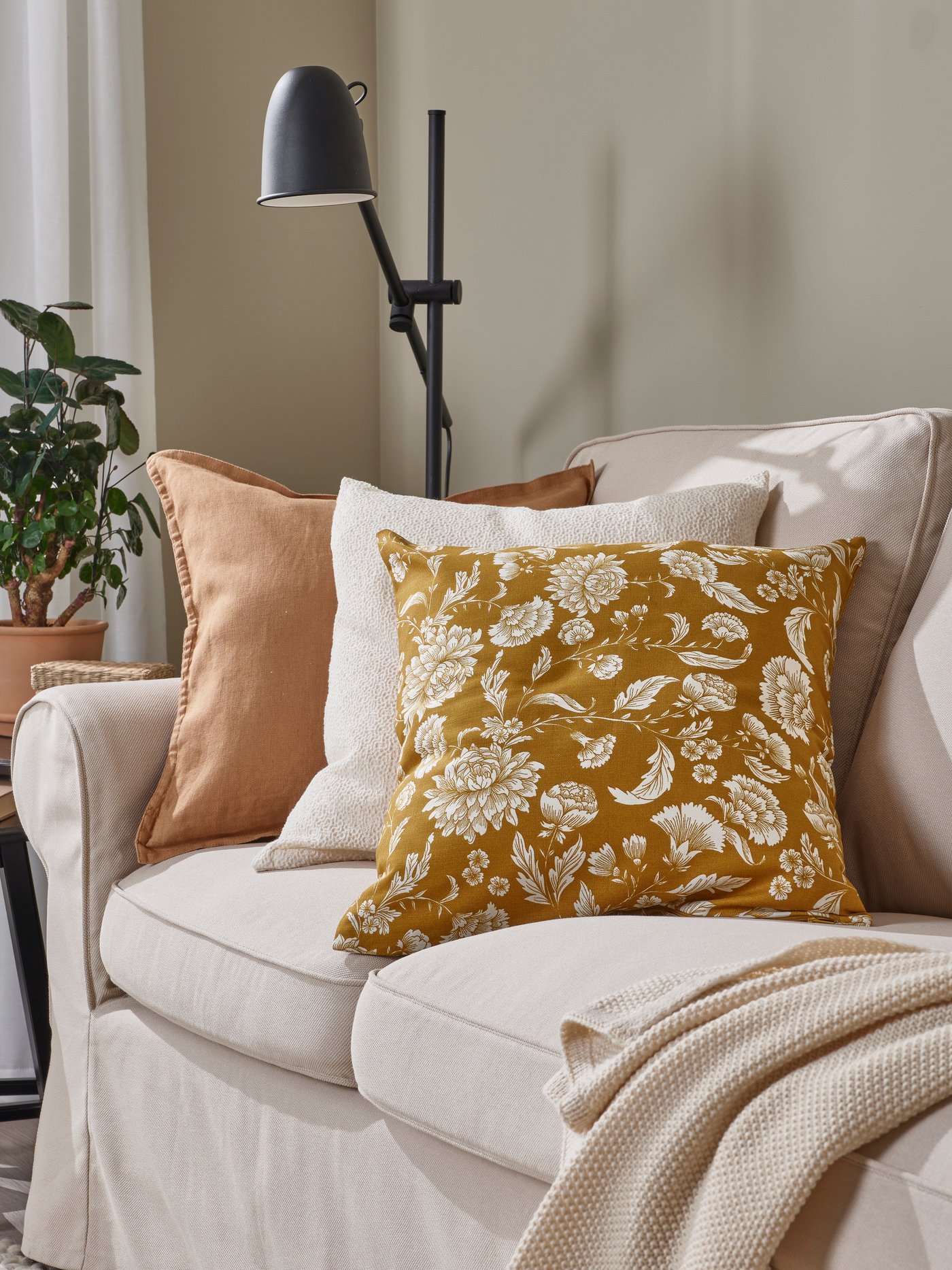 Een beige bank met drie sierkussens waarvan een geelbruine IDALINNEA kussenhoes.