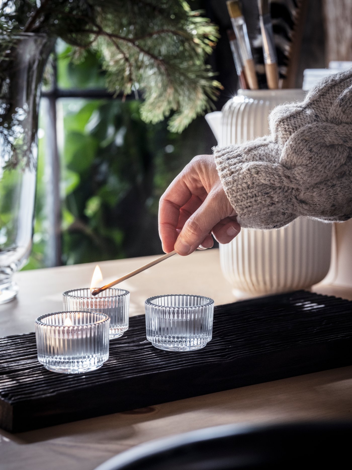 Eine Person zündet mit einem langen Streichholz drei ENSTAKA Duftteelichter an, die zusammen mit einer Vase auf einem Tisch stehen.