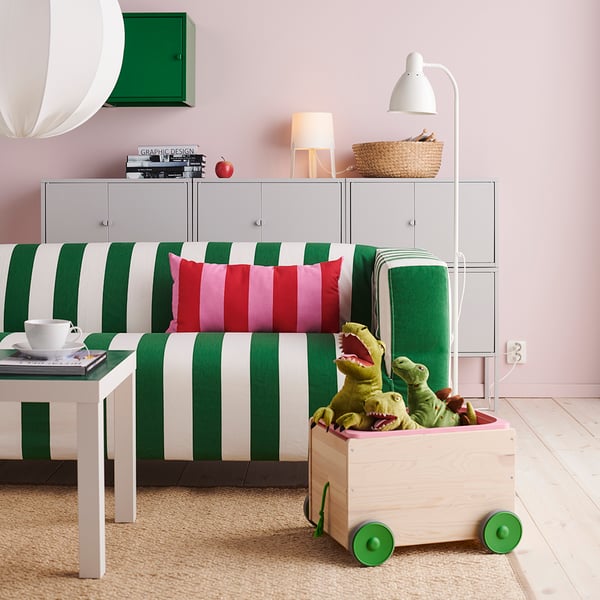 Un carro de madera FLISAT para juguetes con peluches de dinosaurio, en un living con alfombra beige, un sillón KLIPPAN con tapiz a rayas verdes y blancas, una mesa de centro blanca LACK.