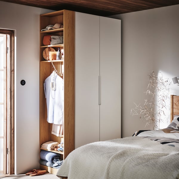 Un armadio modulare in legno chiaro e ante bianche si trova in una camera da letto, con una sezione aperta che mostra vestiti appesi, ripiani con oggetti e coperte piegate, accanto a un letto con una coperta chiara.