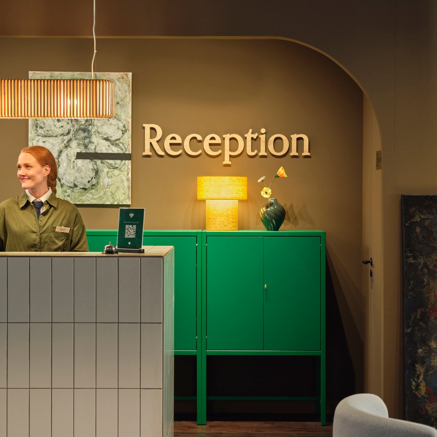 Hotellreception med kaklad disk, personal bakom skrivbord, grönt skåp, lampa och stor "Reception"-skylt på varmfärgad vägg.