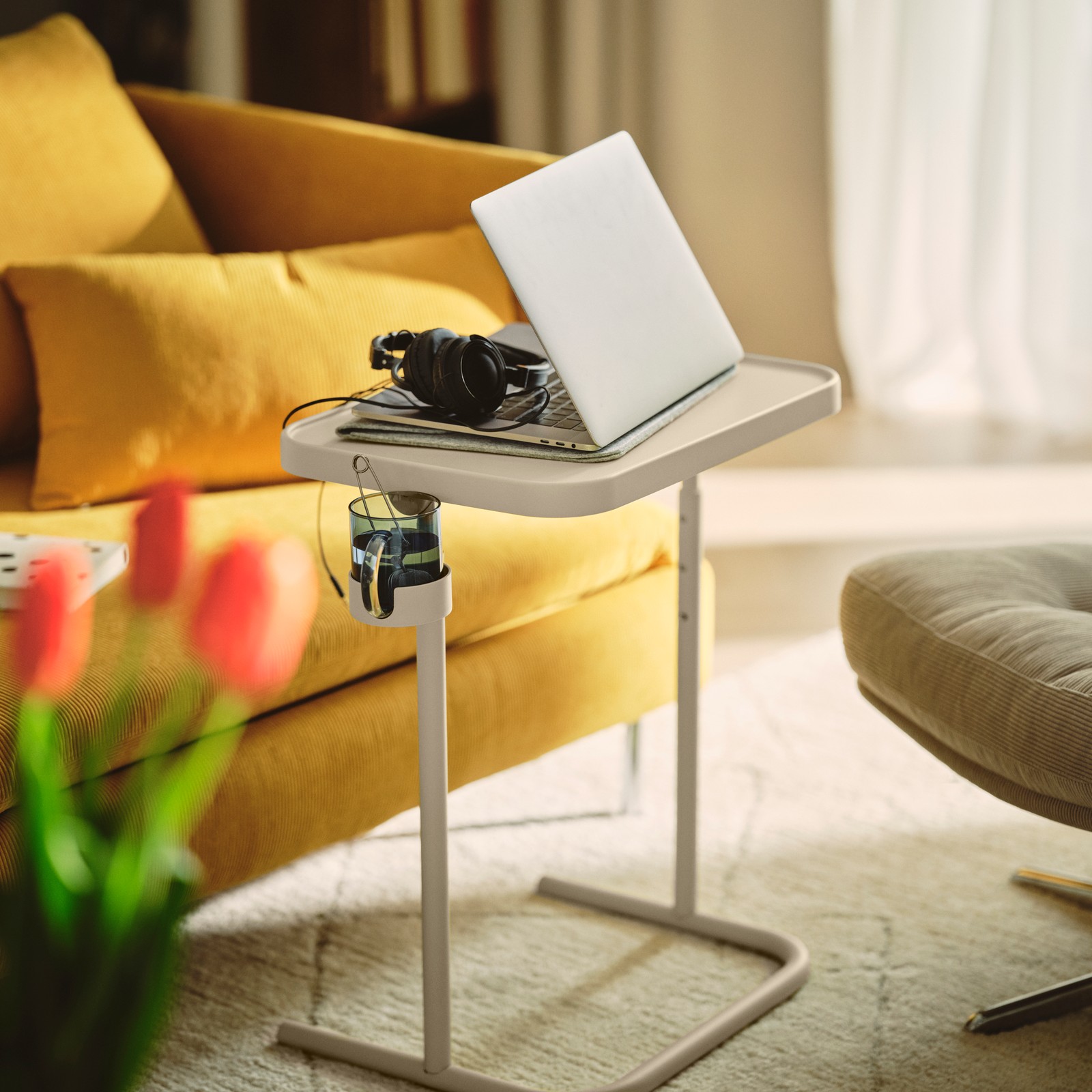 A living room featuring a beige BJÖRKÅSEN laptop stand on a sofa, resting under a laptop and headphones.