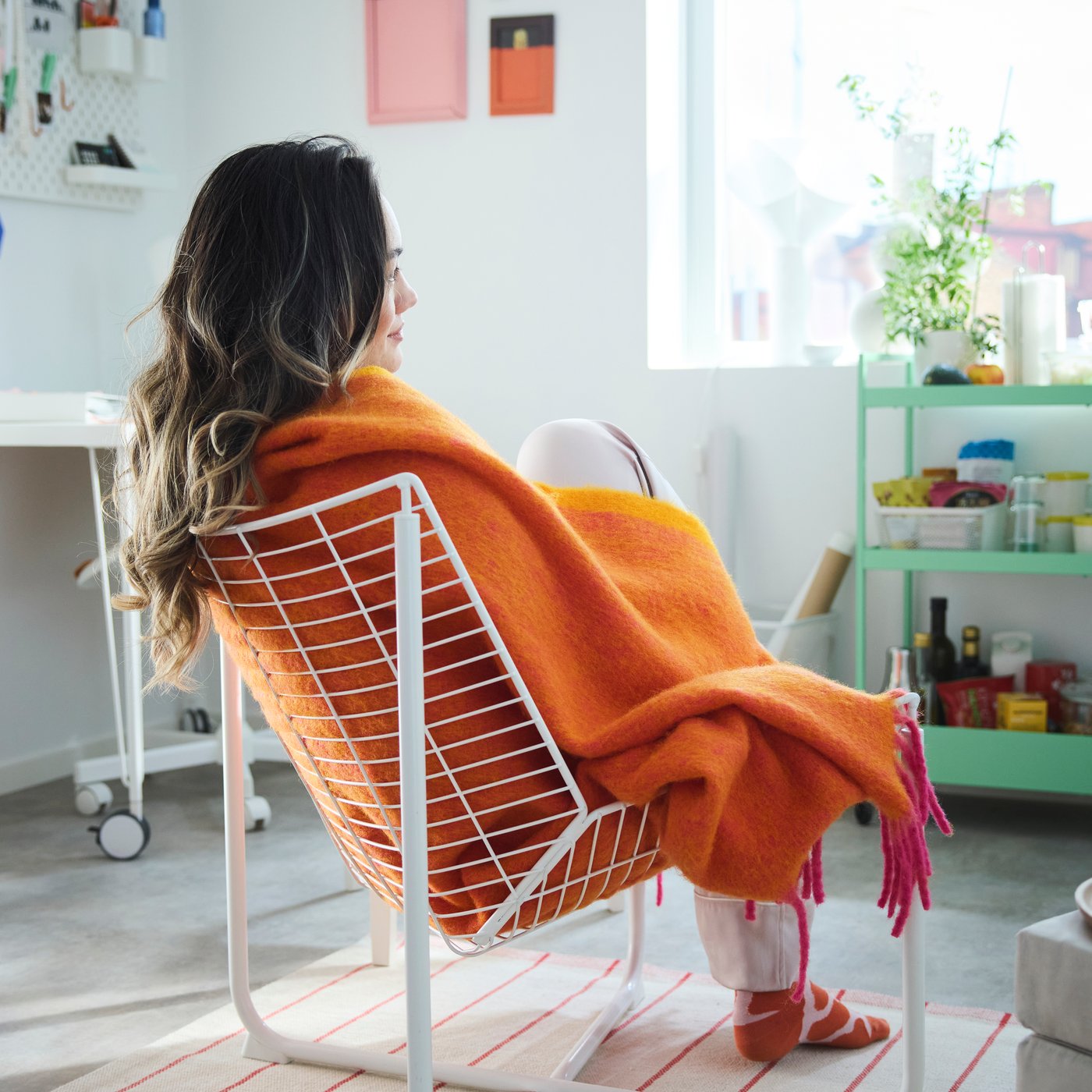 A person sitting in a white net chair wrapped in the orange KLIPPOXEL throw placed in a white room with colourful objects.