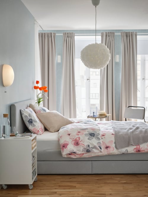 The side of a GLADSTAD bed in front of a curtained window covered with flower pattern bed linen, with a pendant lamp above.
