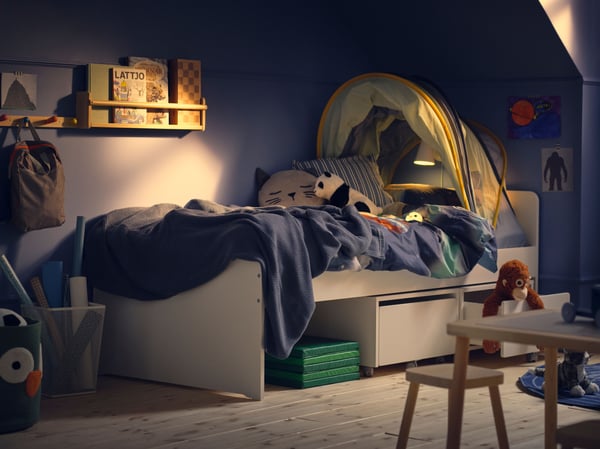 The corner of a children’s bedroom with blue wall in evening light, a SLÄKT bed with blue bedding and a bed tent.