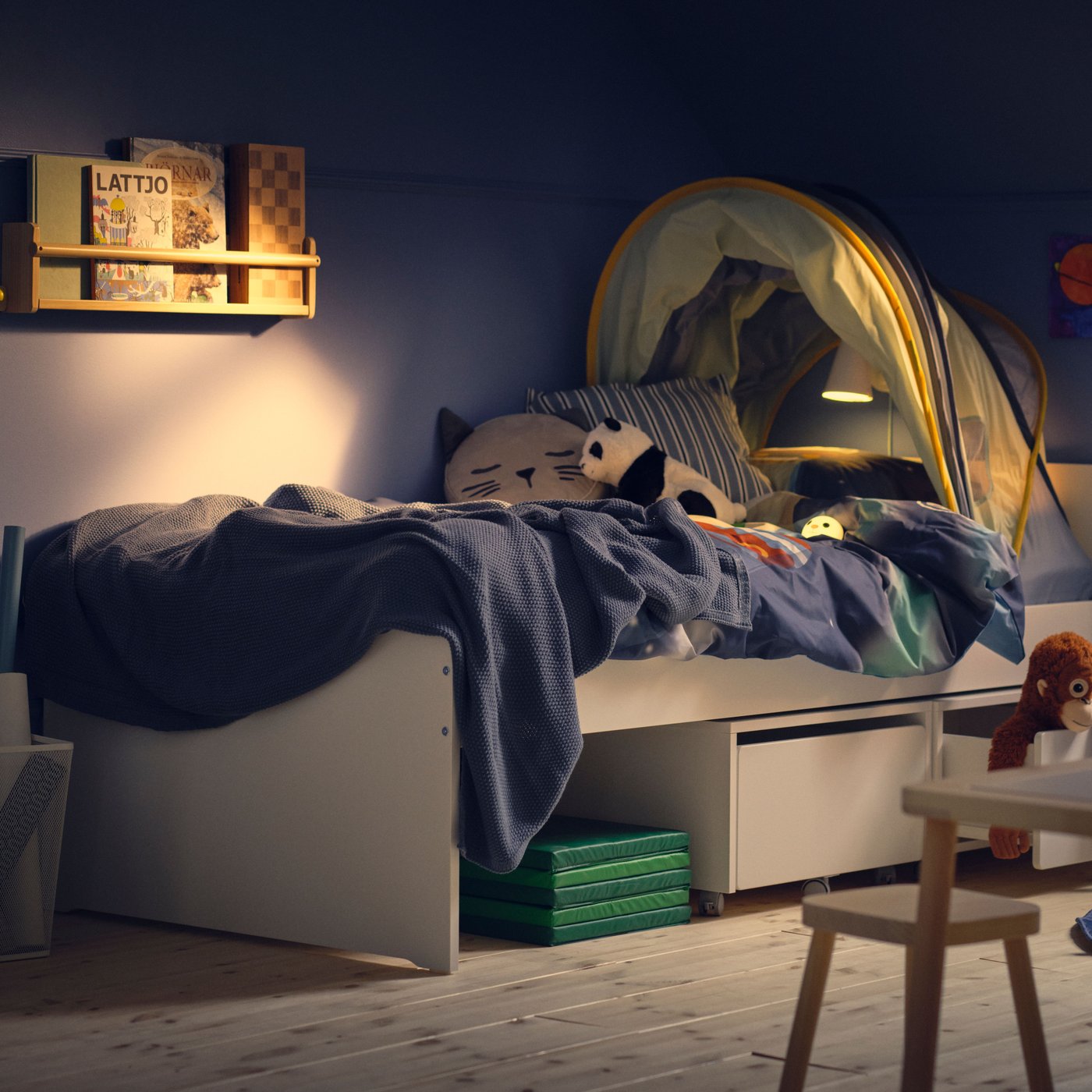 The corner of a children’s bedroom with blue wall in evening light, a SLÄKT bed with blue bedding and a bed tent.