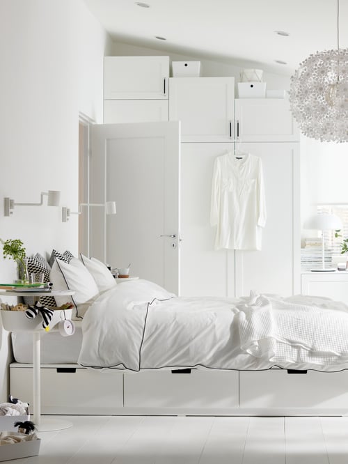 Cadre de lit NORDLI dans une chambre lumineuse avec housse de couette et taie d’oreiller blanches et une suspension blanche aérienne.