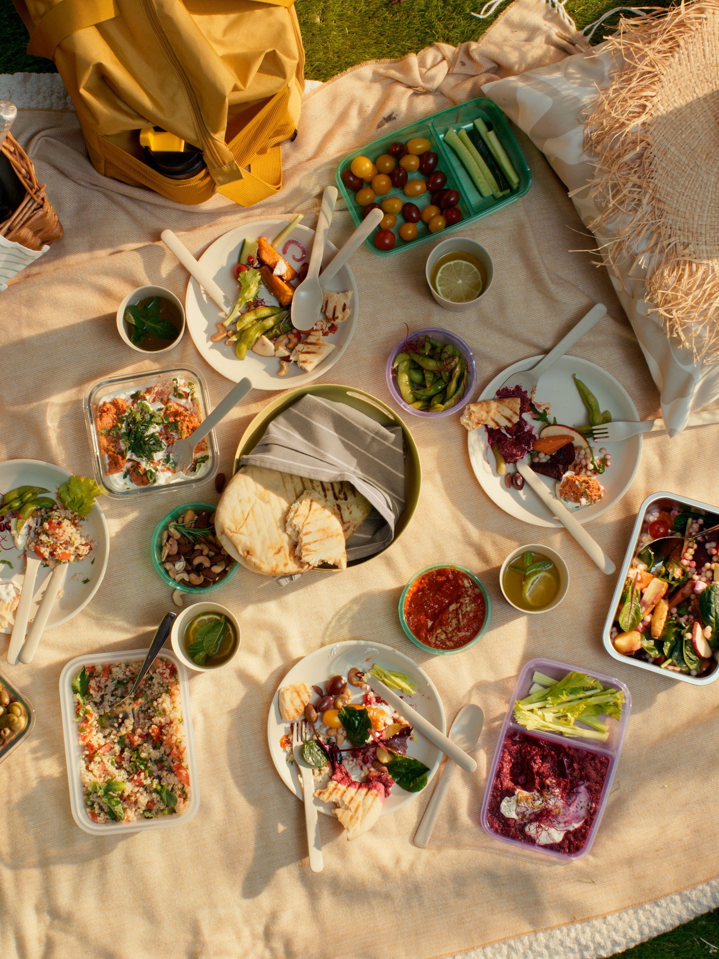 Sommerliches Picknick auf einer hellen Decke mit verschiedenen frischen Speisen, darunter Salate, Gemüse, Brot und Dips. Mehrere Schalen, Teller und Getränke sind dekorativ arrangiert, umgeben von einer gelben Tasche und einem Strohhut im Sonnenlicht.