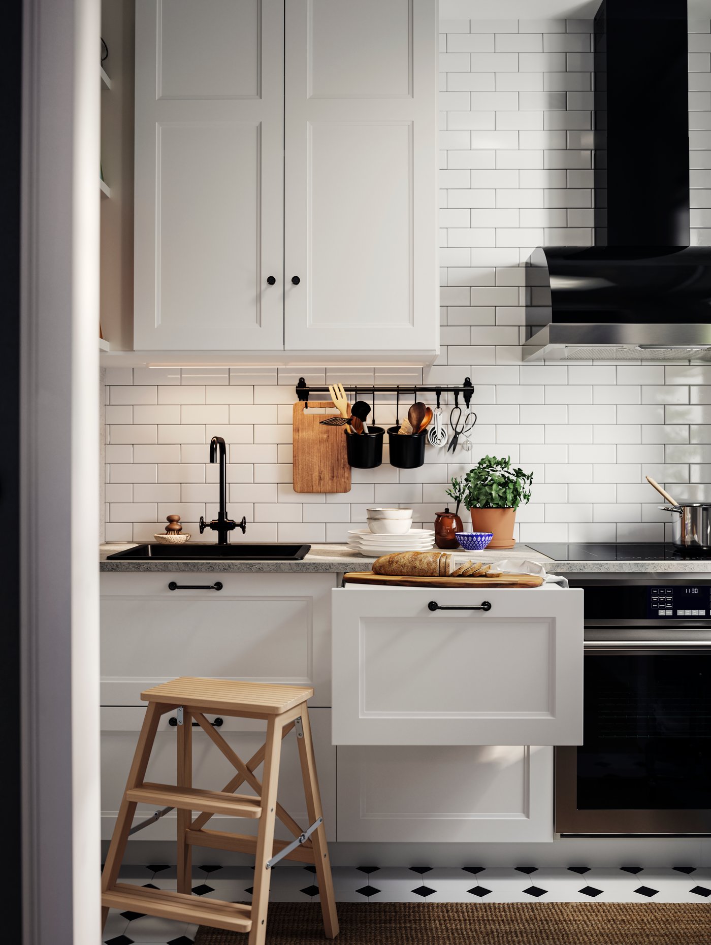 매트 화이트 AXSTAD 악스타드 주방 앞판, 스텝스툴, 블랙 손잡이가 보이고 조리대 위에 빵, 접시, 화초가 담긴 화분이 놓인 모습.