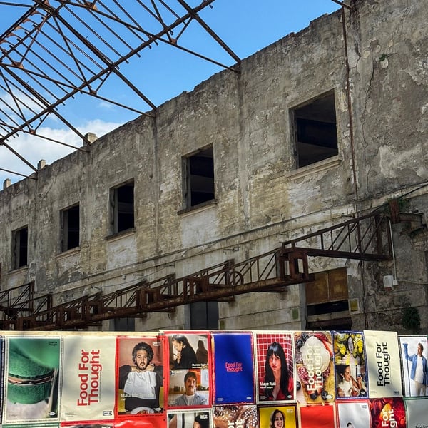 Facciata di edificio industriale dismesso con struttura del tetto a vista, sopra una lunga fila di poster colorati “Food For Thought” affissi in basso.
