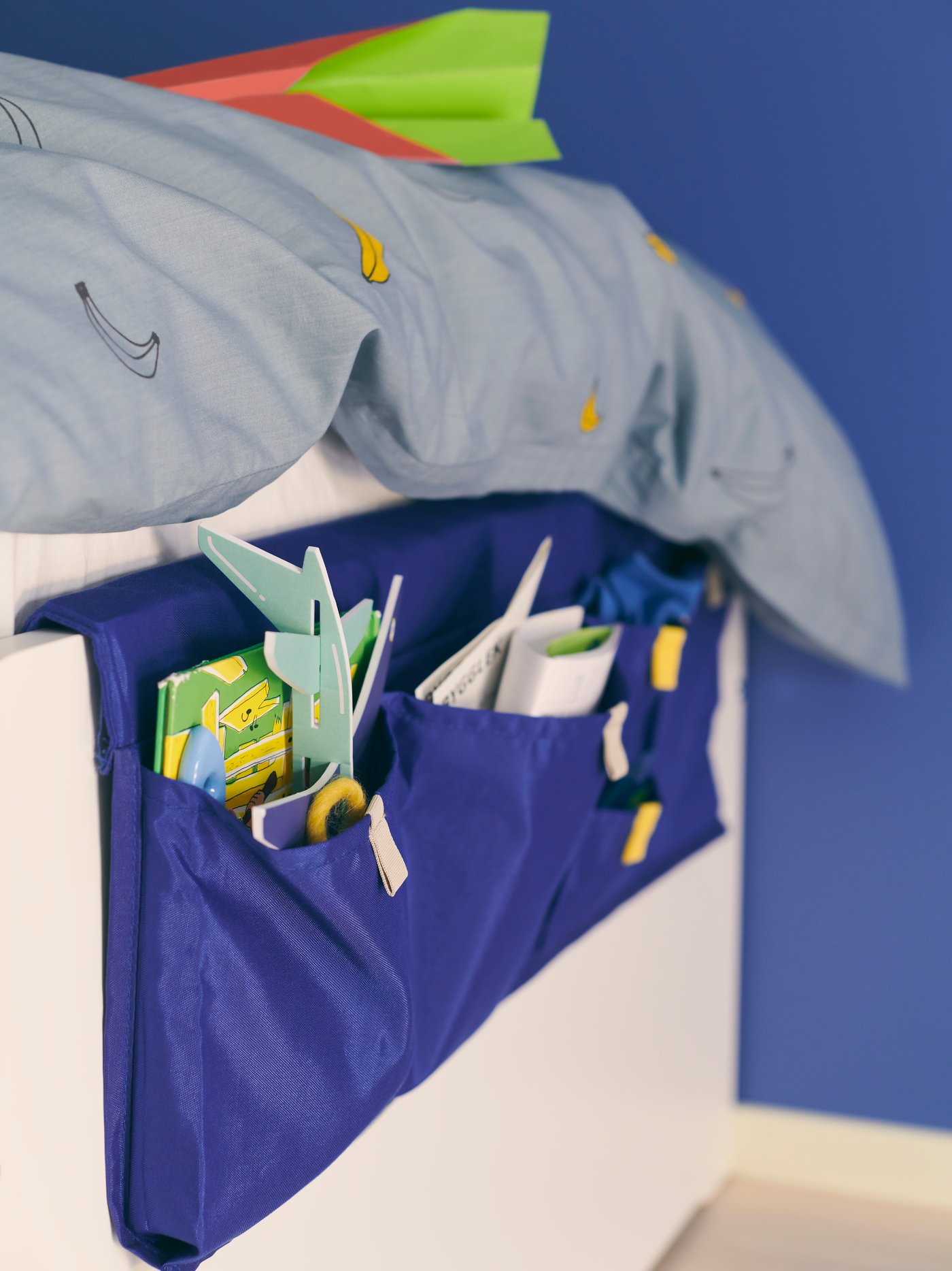 Una solución de almacenaje con bolsillos para la cama MÖJLIGHET colocada a los pies de una cama blanca. La cama está junto a una pared azul.