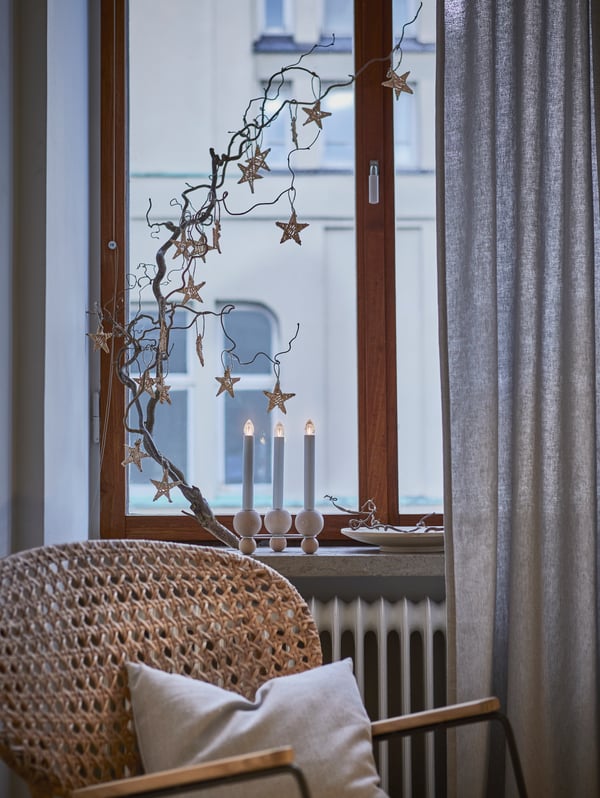 Beech STRÅLA LED three-armed candelabra is on a windowsill beneath a branch with VINTER 2021 hanging star decorations.