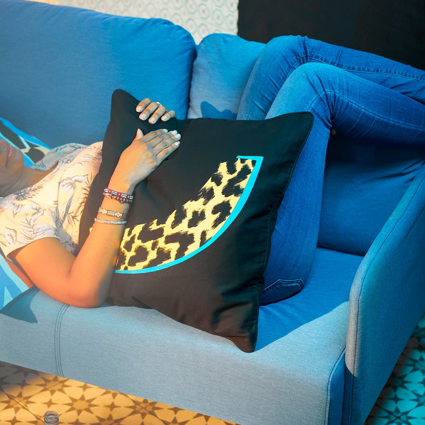 Seseorang berseluar jeans berbaring di sofa berwarna biru firus, memeluk bantal besar dengan sarung kusyen ÖMSESIDIG berwarna hitam/kuning.