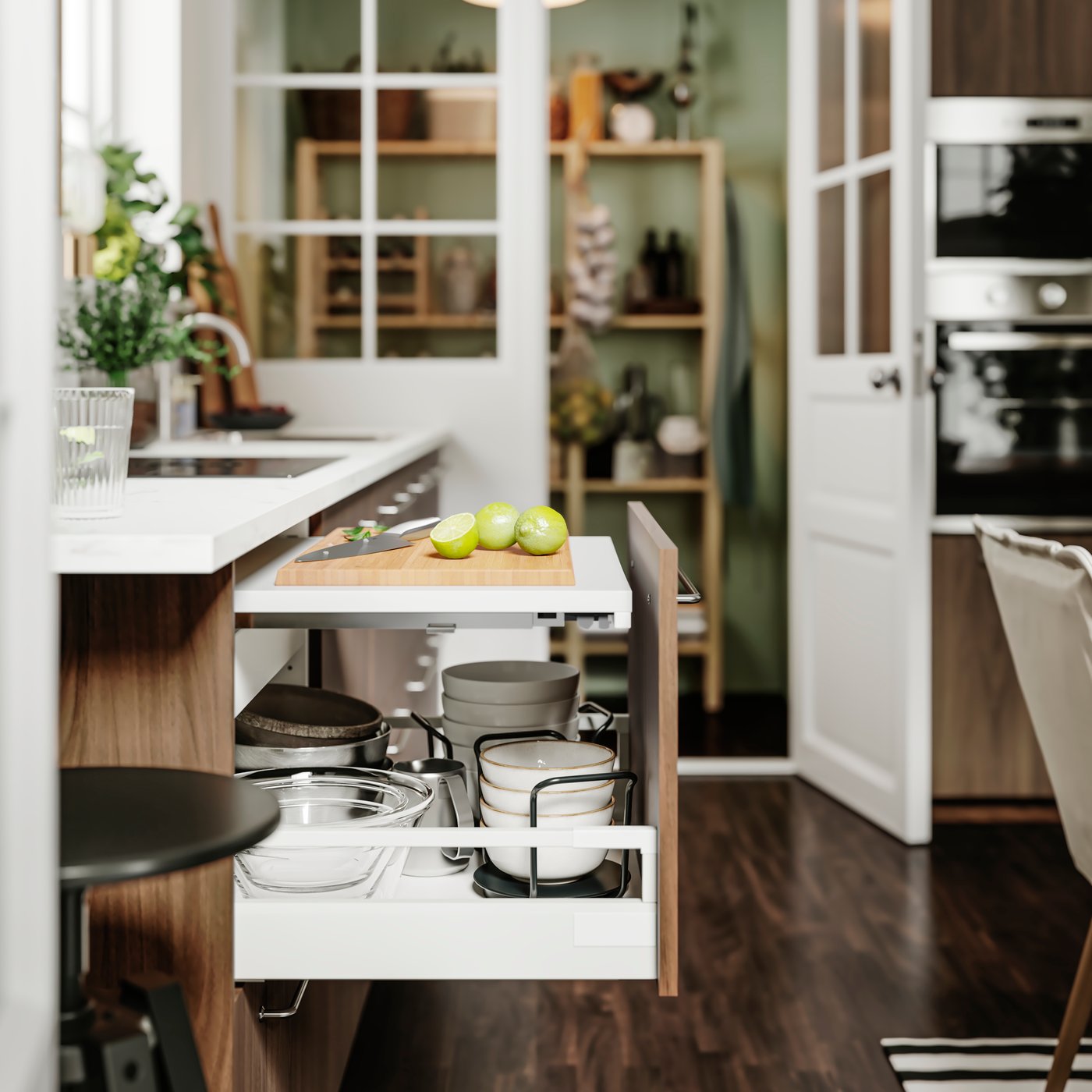 Ett ljust kök med öppna vitrindörrar som visar hyllor med porslin och köksredskap; i förgrunden syns en köksö i trä med pallar och gröna äpplen, mörkt trägolv och dagsljus som flödar in.
