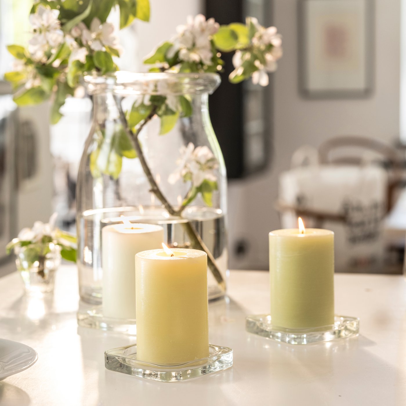 Zapálené svíčky IKEA na skleněných podložkách vedle vázy s větvičkou kvetoucího stromu, aranžované na světlém stole v moderním interiéru.