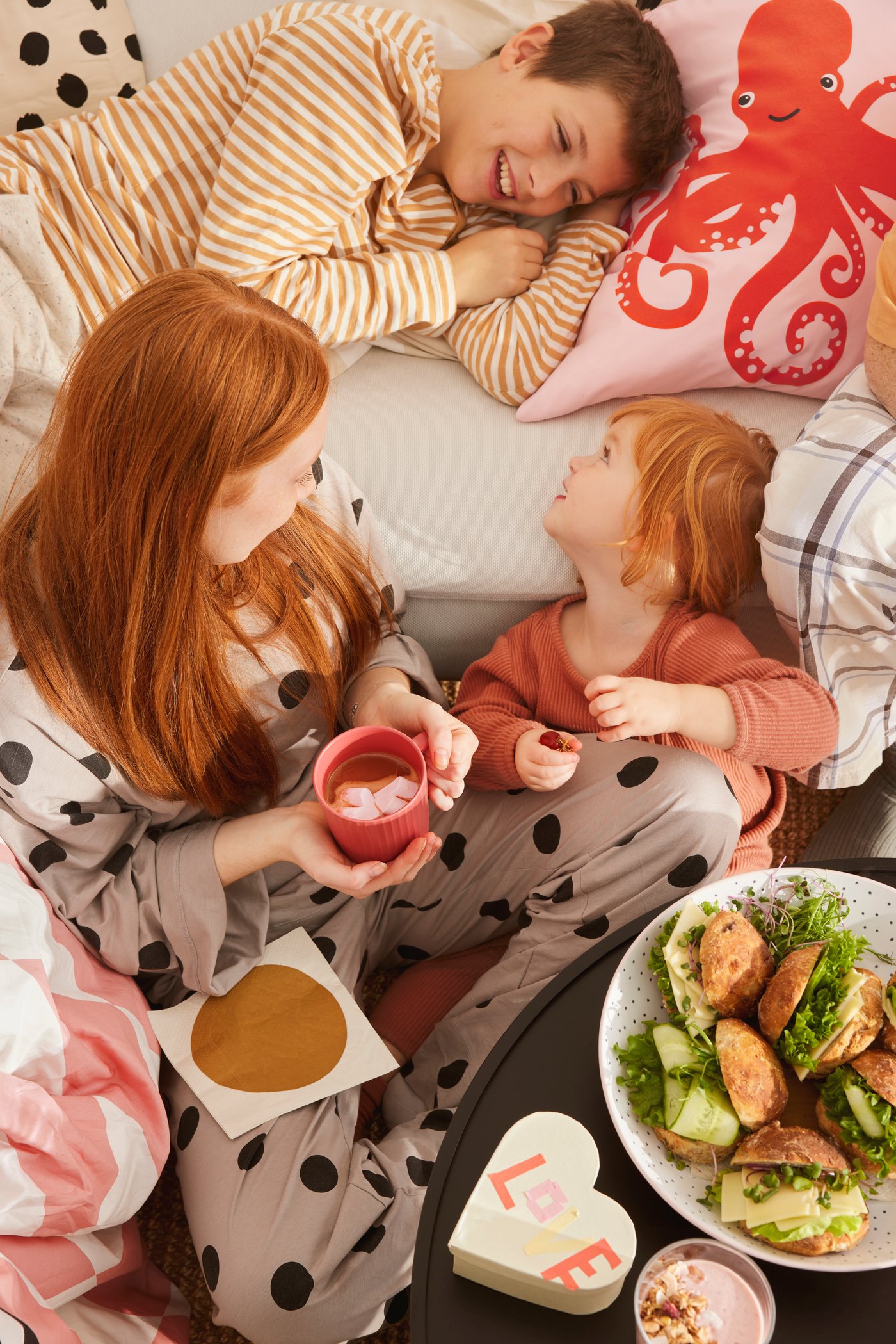 En mor sidder sammen med hendes to børn og drikker kaffe ved sengen med puder. 