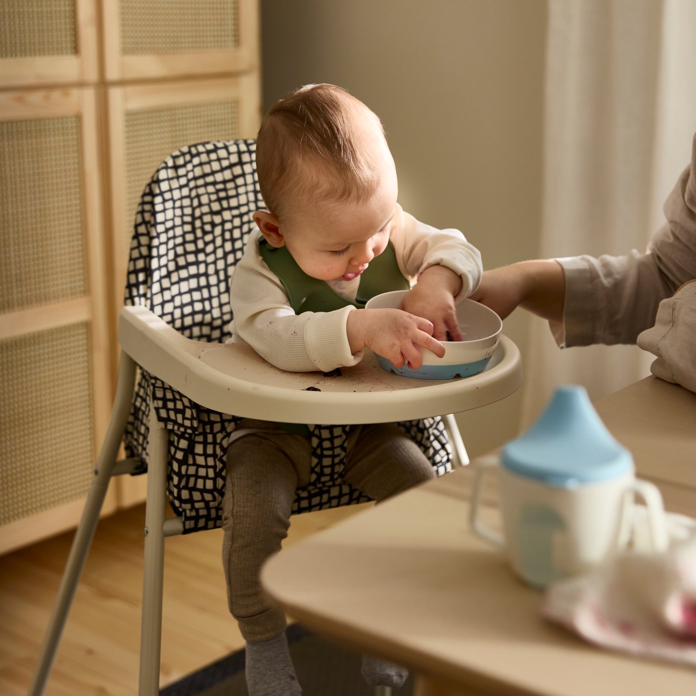 Ein Baby sitzt in einem SANDKATT Kinderhochstuhl mit Tablett und spielt gerade mit einer BÖRJA Schale. Daneben steht eine erwachsene Person und passt auf.