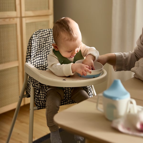 A baby sitting in the SANDKATT highchair playing with a BÖRJA plate/bowl next to an adult supervising.