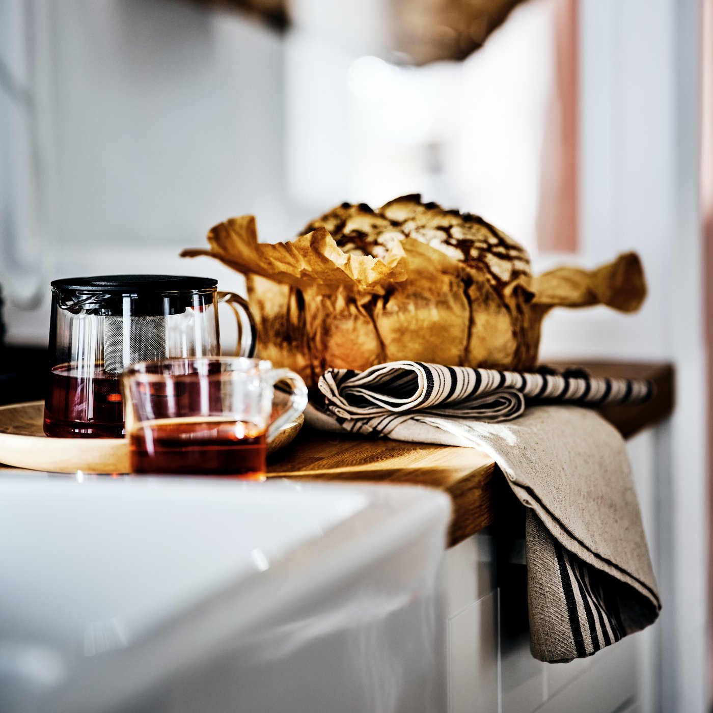 A white kitchen cabinet with a wood counter holding an IKEA 365+ mug and RIKLIG teapot filled with tea, beside a large cake.