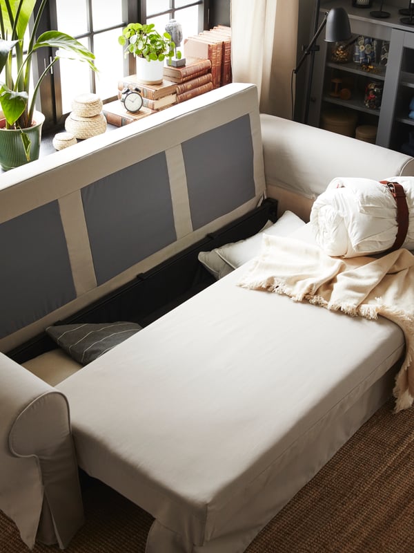 A light beige sofa-bed is being converted into a sofa. Pillows and cushions lie in the storage space under the seat.
