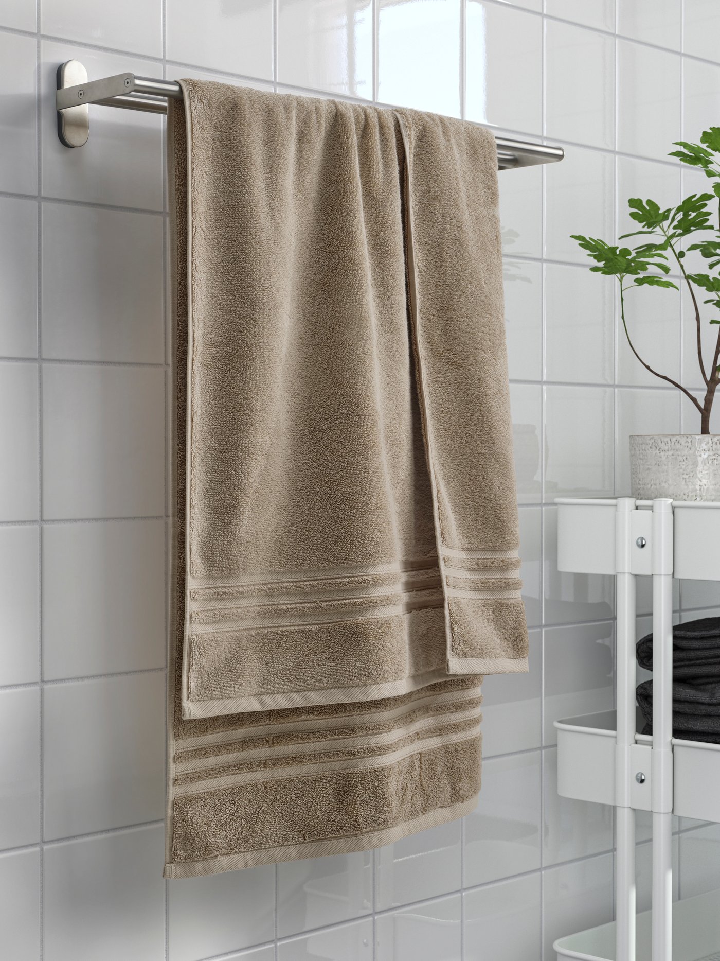 タイル張りのバスルームで、金属製タオルラックに掛けられたベージュのタオルと、近くの白い棚に置かれた小さな鉢植えと畳まれたタオル。