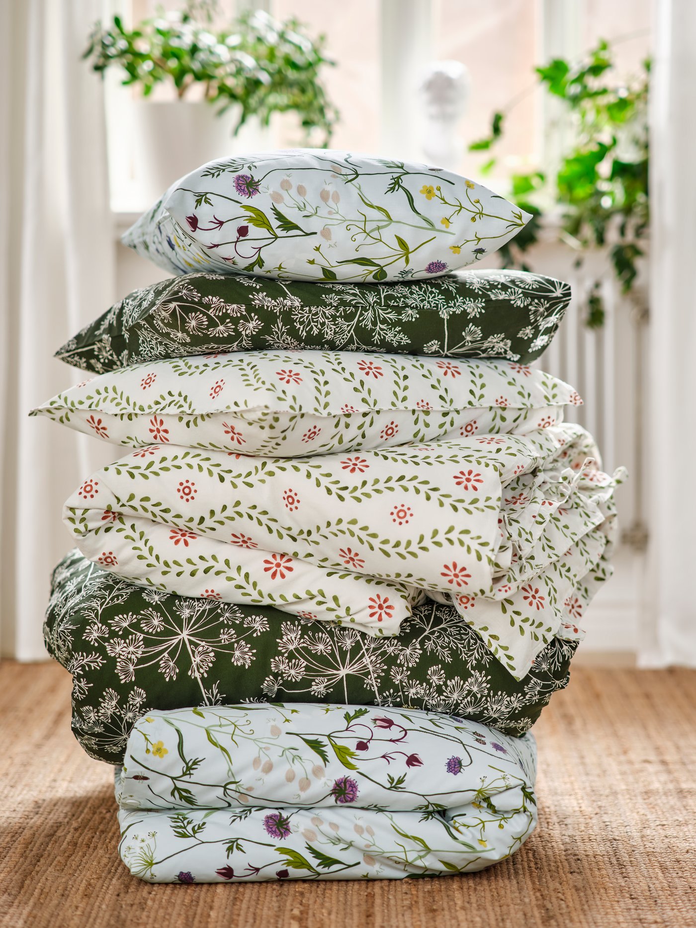 A bedroom showcasing a stack of FYRISVIDE, MATTOXBÄR and DAGGBJÖRNBÄR duvet covers and pillowcases on a jute rug.