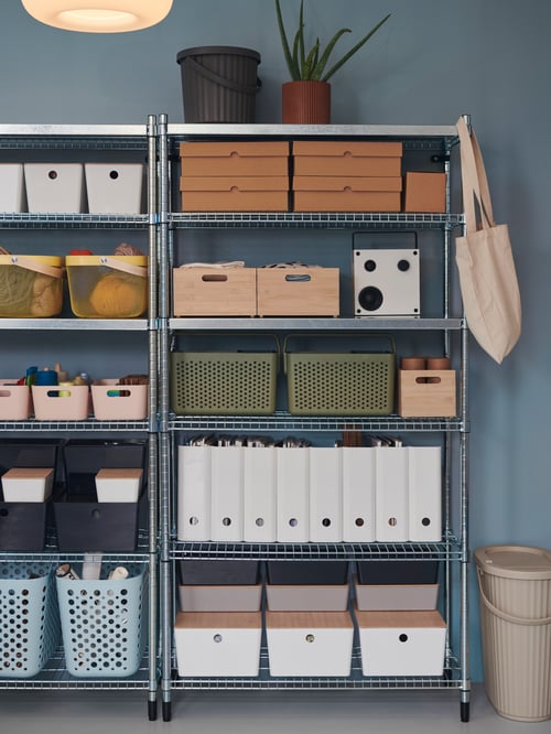 In a hallway, there is a shelf section with KUGGIS boxes with lids, storage baskets, magazine files and a black bin with lid.