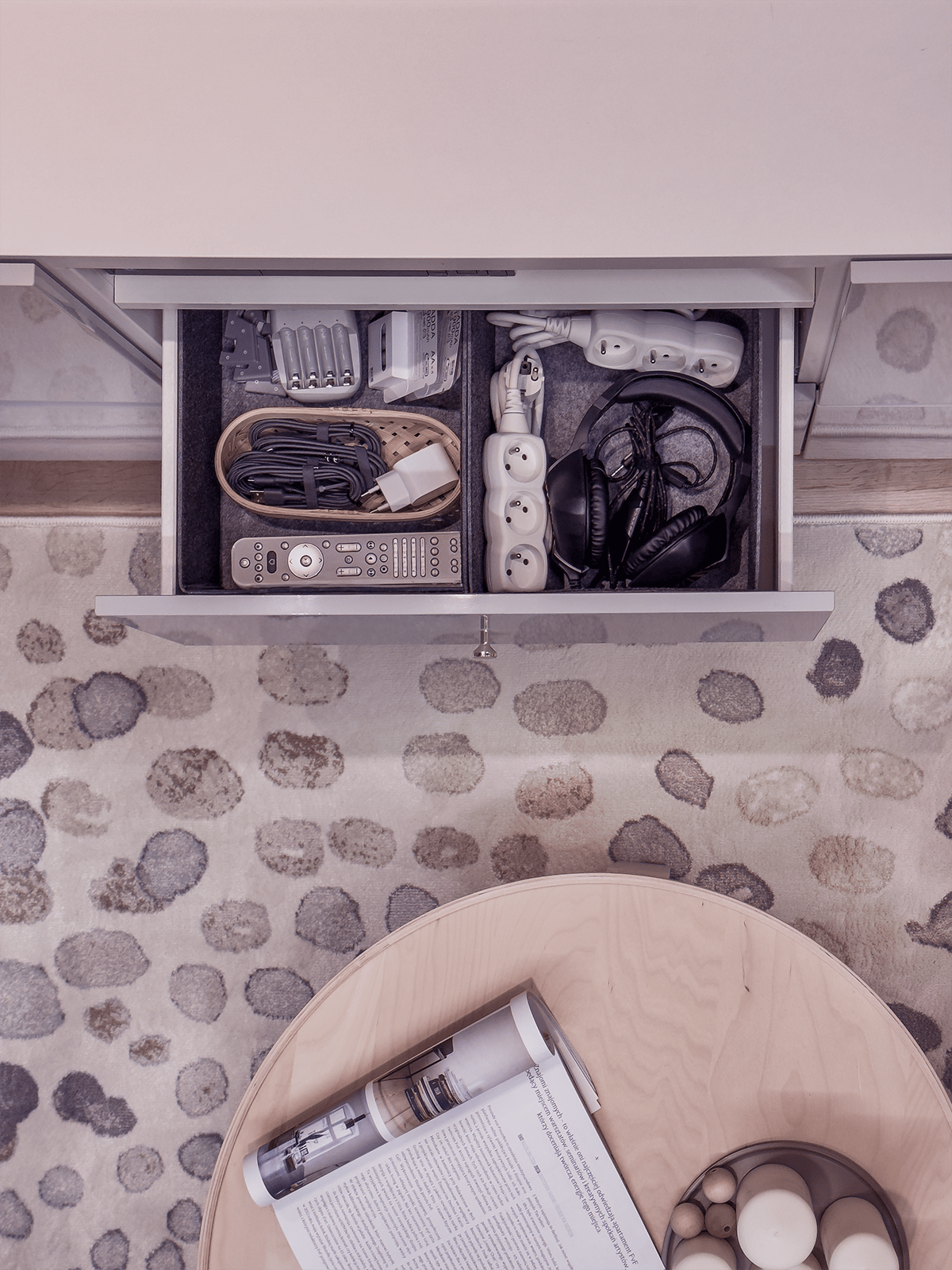 An open drawer in a sideboard filled with organised electronic items, including remote controls, cables, and power strips. The photo is taken from above and also shows a round wooden table and a light rug with a stone‑like pattern.